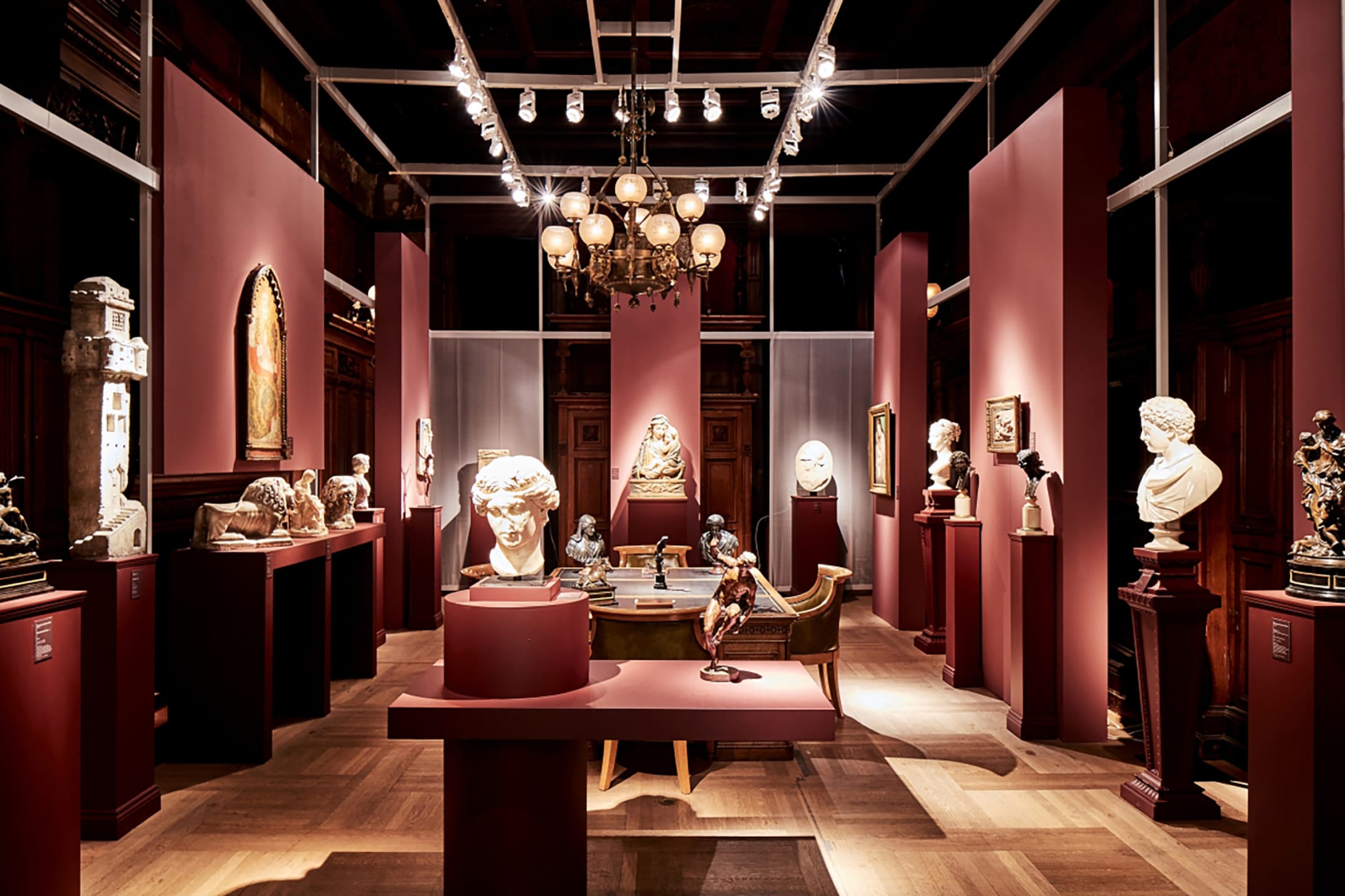 Art gallery interior with various sculptures and paintings displayed on red walls under elegant lighting.