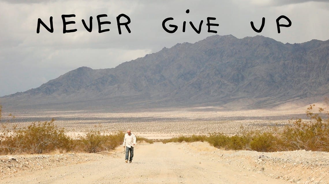 Man walking alone in a desert landscape with mountains in the background, captioned "Never Give Up" in large black letters.