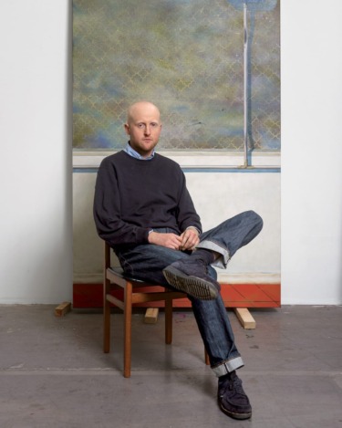 Person sitting on a chair with a painting in the background, wearing a black sweater and jeans, looking at the camera.