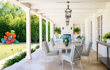 Spacious outdoor patio with white table, blue chairs, hanging lanterns, potted plants, and colorful sculpture in garden.