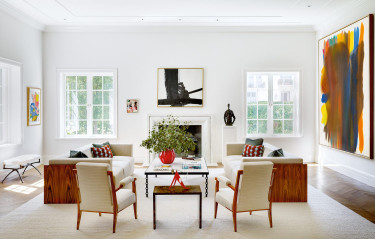 Modern living room with white sofas, colorful art, large windows, and a central coffee table with a red vase.