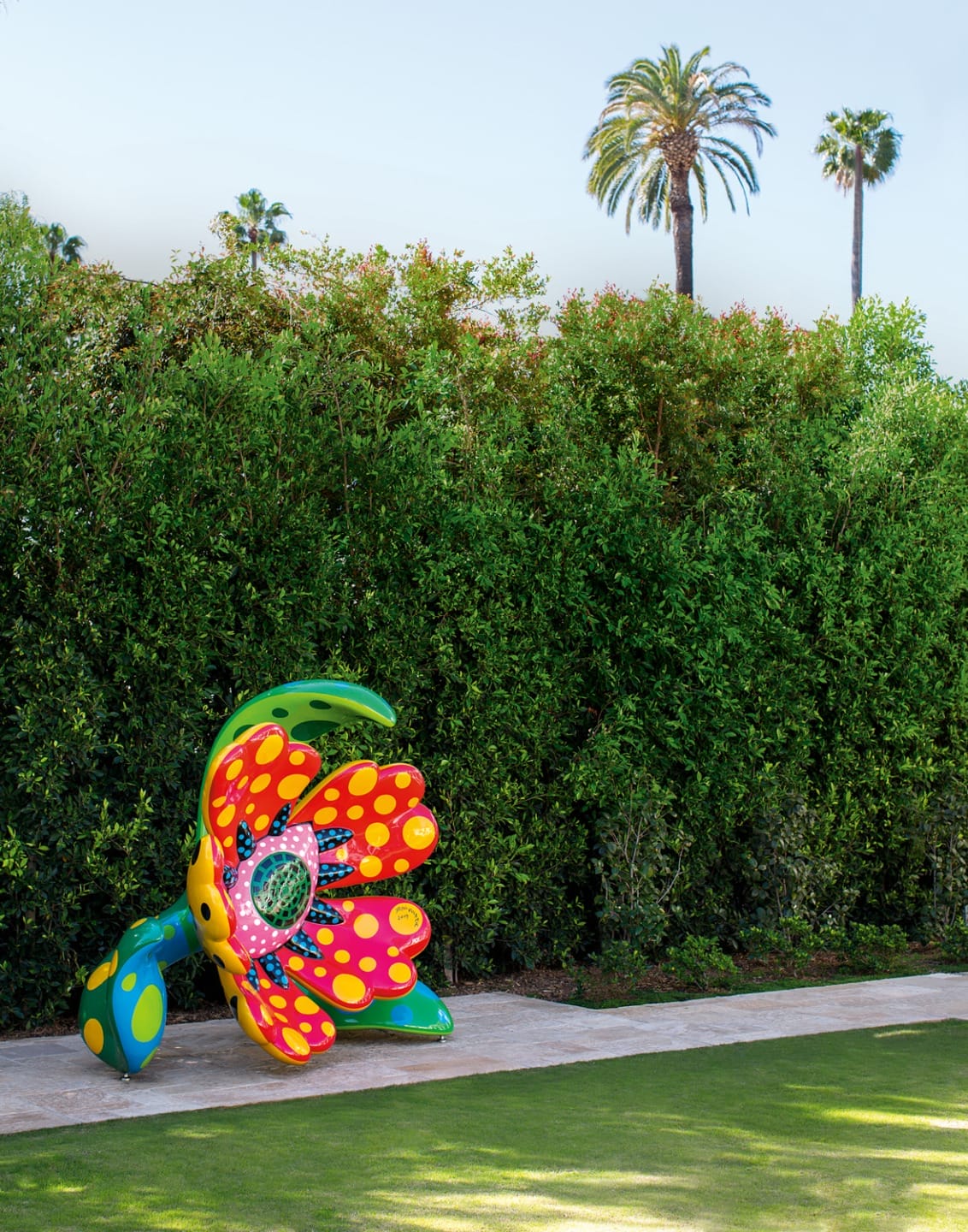 Colorful flower sculpture with polka dots beside a large hedge on a sunny day, palm trees in the background.