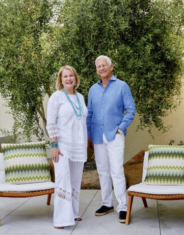 A smiling couple standing outdoors on a patio surrounded by olive trees and lounge chairs with decorative cushions.