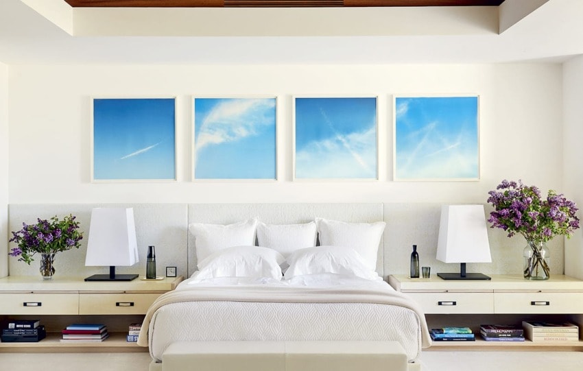 Modern bedroom with a white bed, purple flowers, and four framed blue sky pictures on the wall above the headboard.