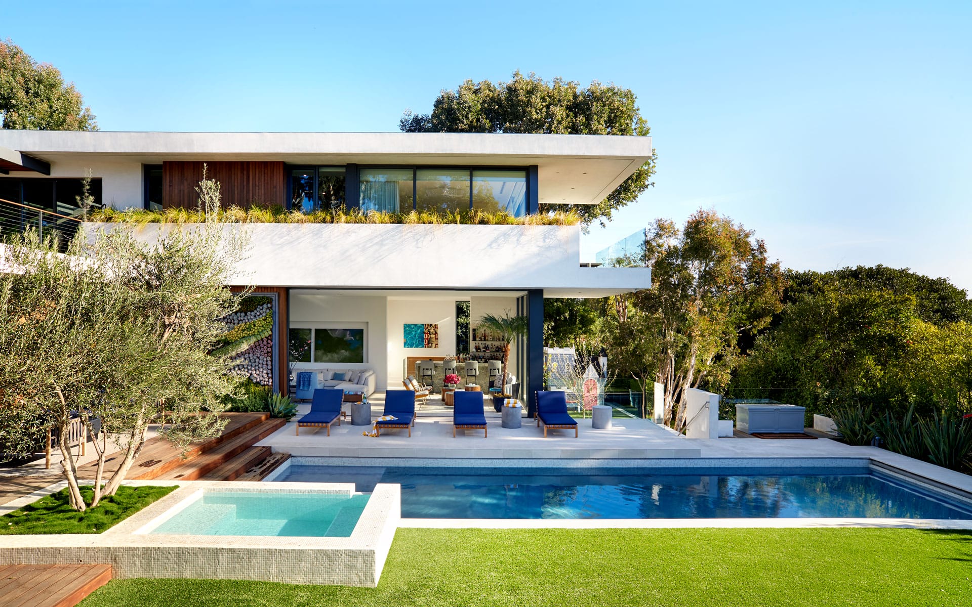 Modern two-story house with a blue pool in the foreground, surrounded by green trees and a clear sky.