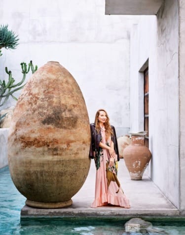 Woman in a pink dress standing next to a large clay pot by a pool with cacti and stone walls in the background.