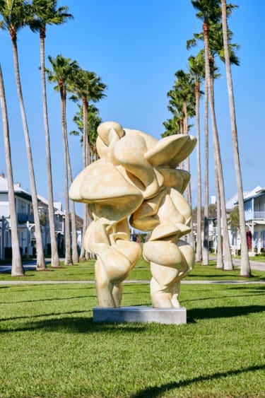 Abstract yellow sculpture on a green lawn with palm trees and houses in the background under a clear blue sky.