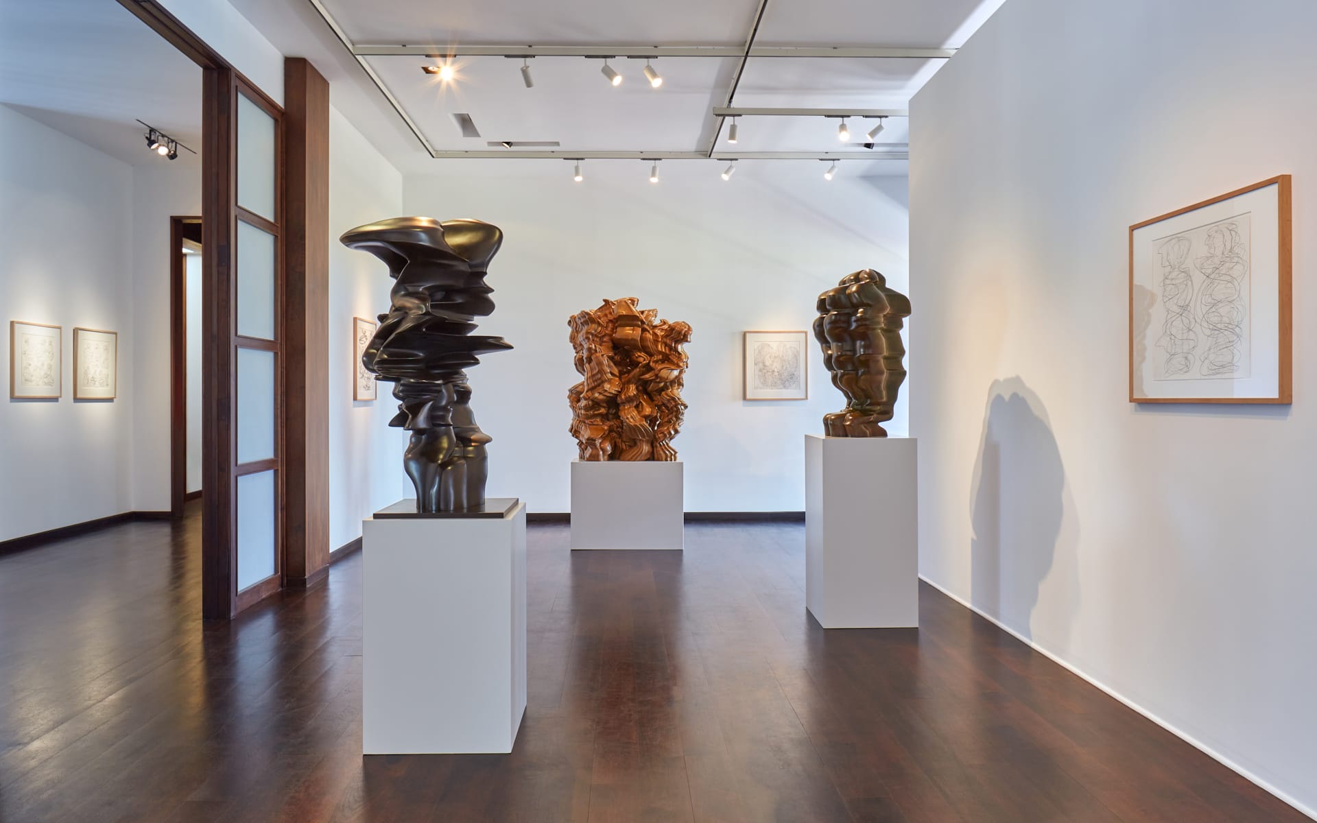 Contemporary art gallery with abstract sculptures on pedestals, framed drawings on walls, and wooden flooring.
