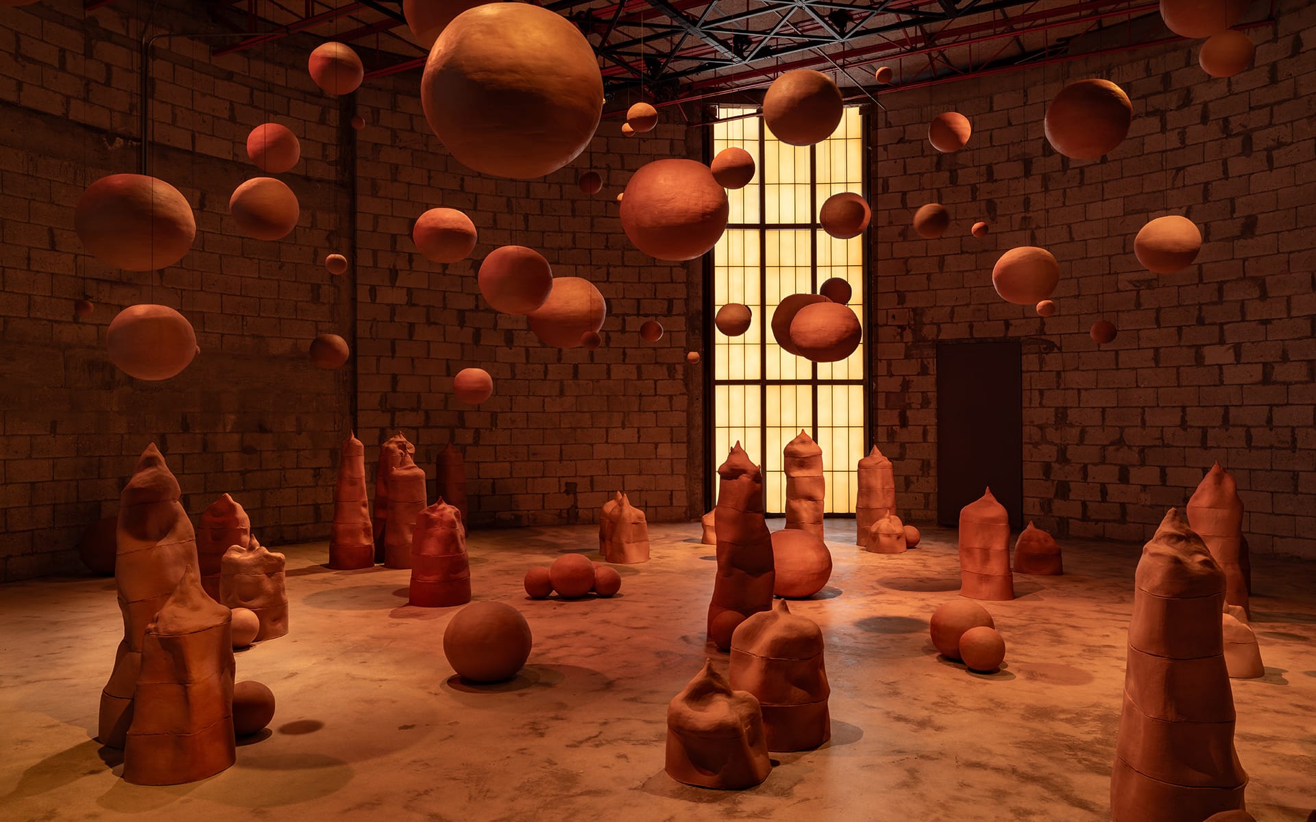 Art installation of suspended terracotta spheres and upright sculptures in a dimly lit room with textured brick walls.