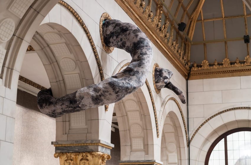 Large snake-like sculpture weaving through ornate arches in a grand room with decorative gold accents and high ceilings.