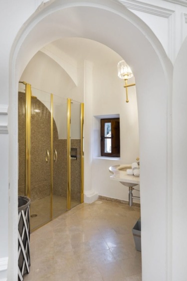 Elegant bathroom with arched entrance, gold-framed shower, small window, and chic sink area.