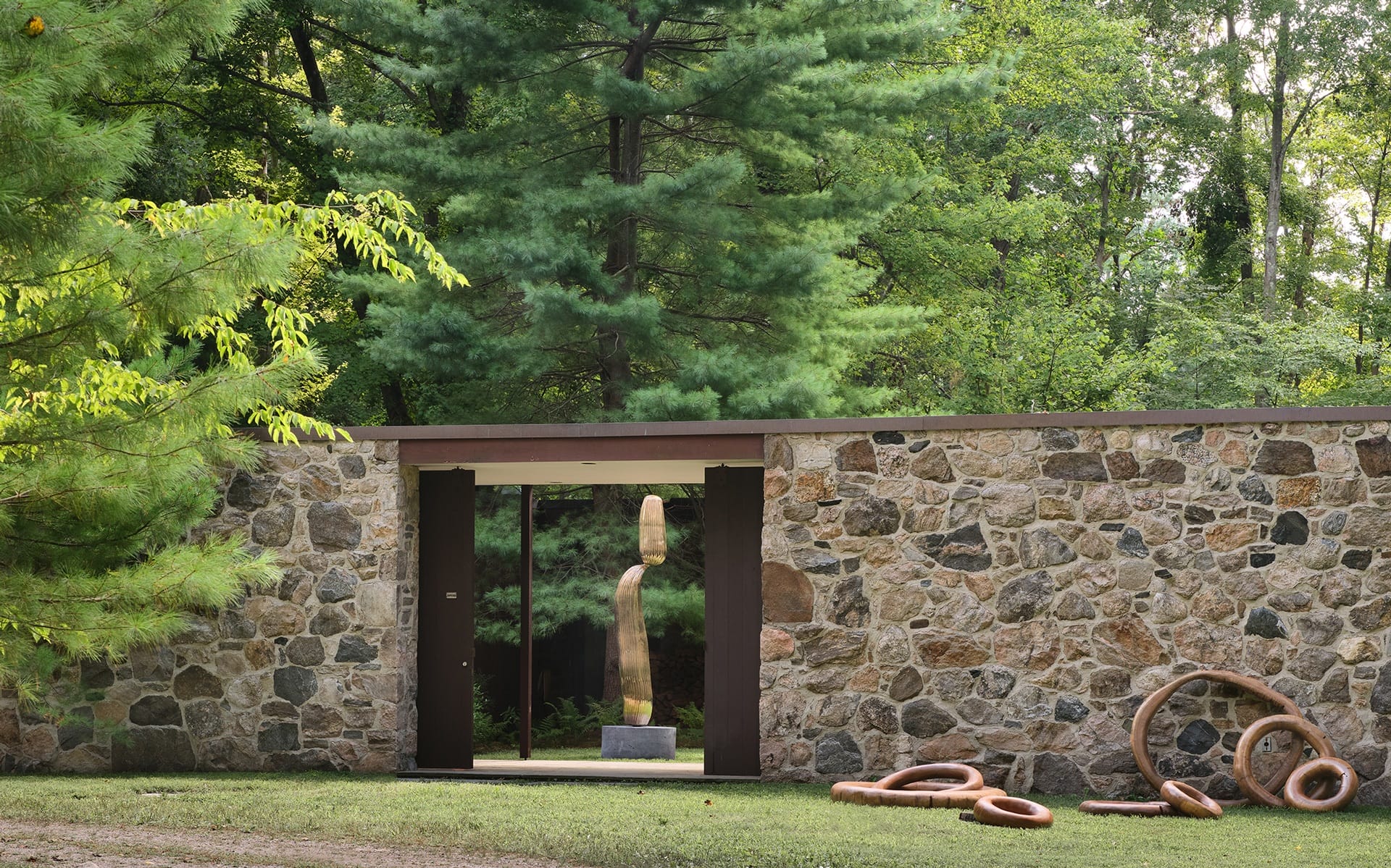 Modern stone building entrance with a tall sculpture inside, set against a backdrop of lush green trees and scattered ring sculptures.