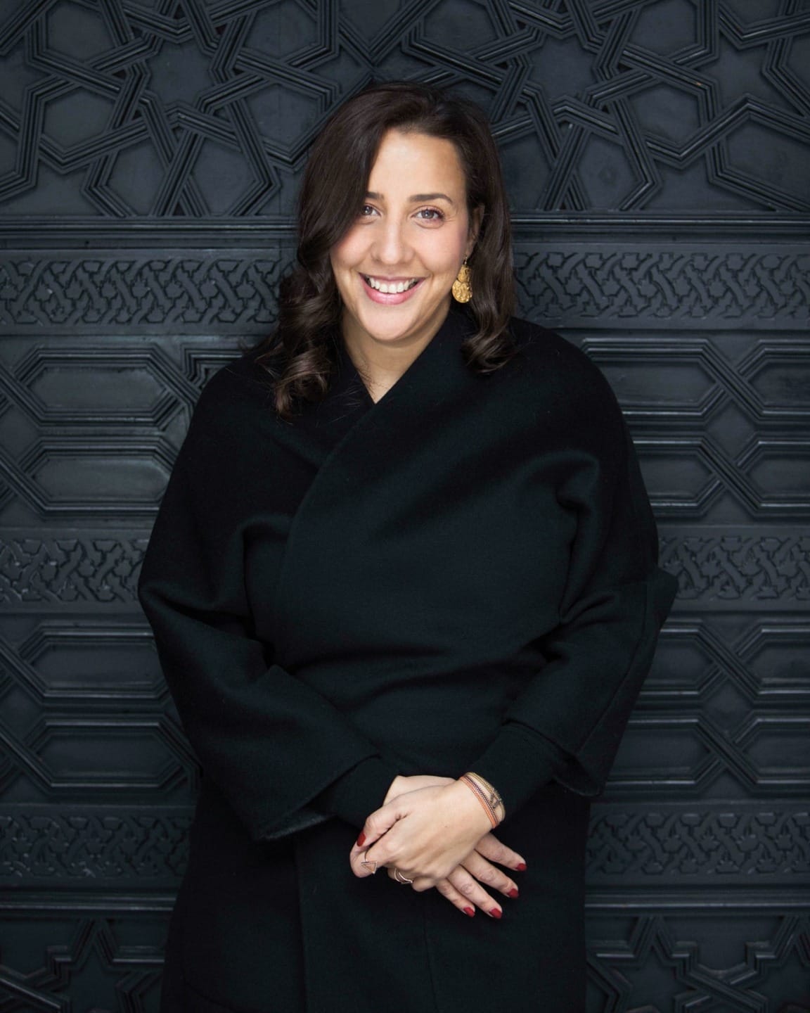 Person smiling in front of a patterned dark wall, wearing a black coat and gold earrings.