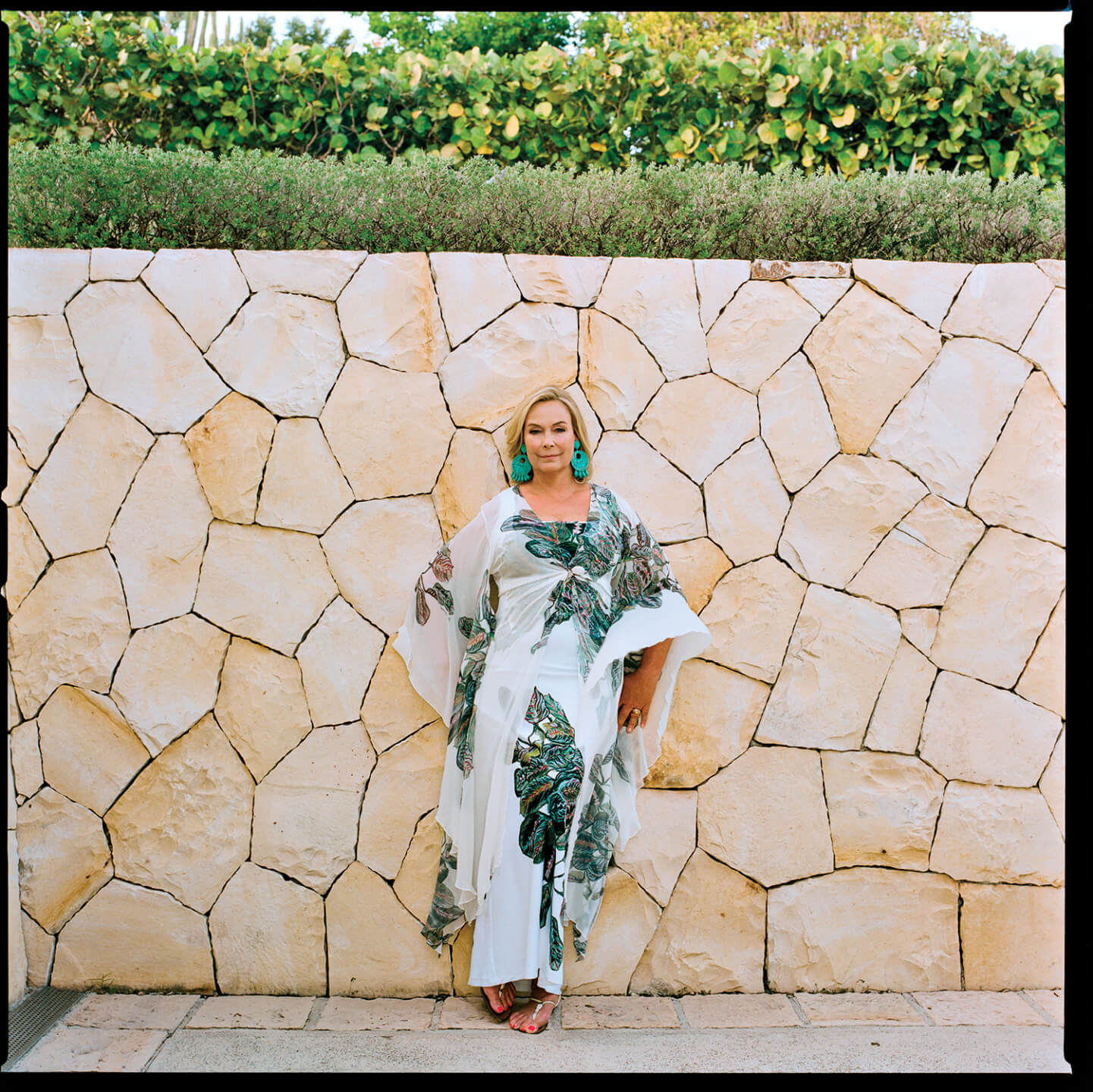 Woman in a white and green leafy dress standing against a stone wall surrounded by green foliage.