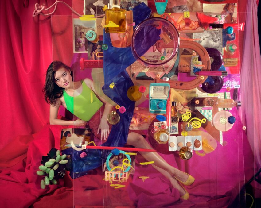 Colorful artistic scene with a woman lying among vibrant objects and fabrics, creating a surreal, layered composition.