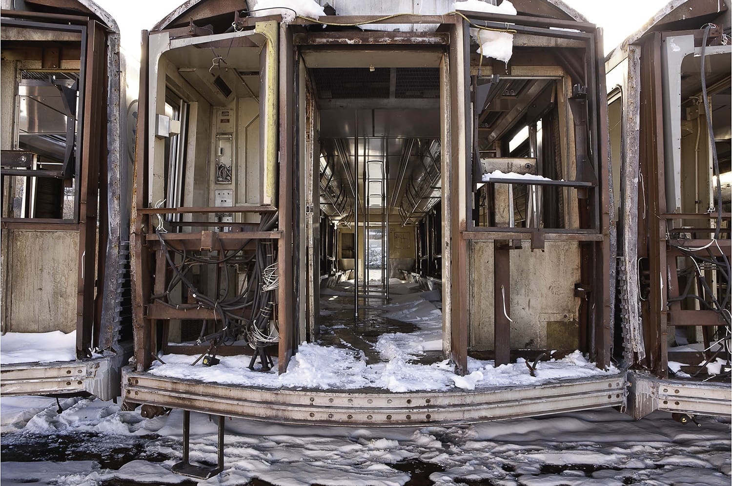 Abandoned train cars with snow-covered floors and open doors, revealing rusted interiors and disused electrical equipment.