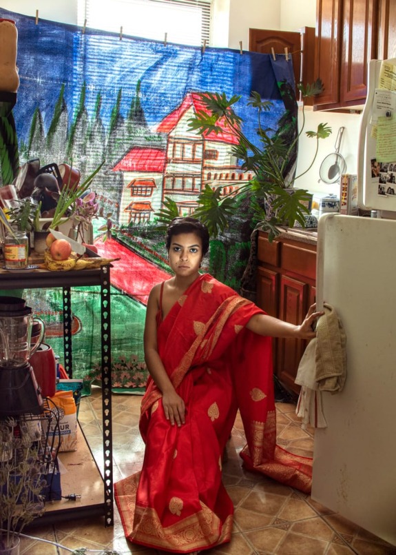 Person in red sari sitting on a kitchen floor with a colorful backdrop behind them.