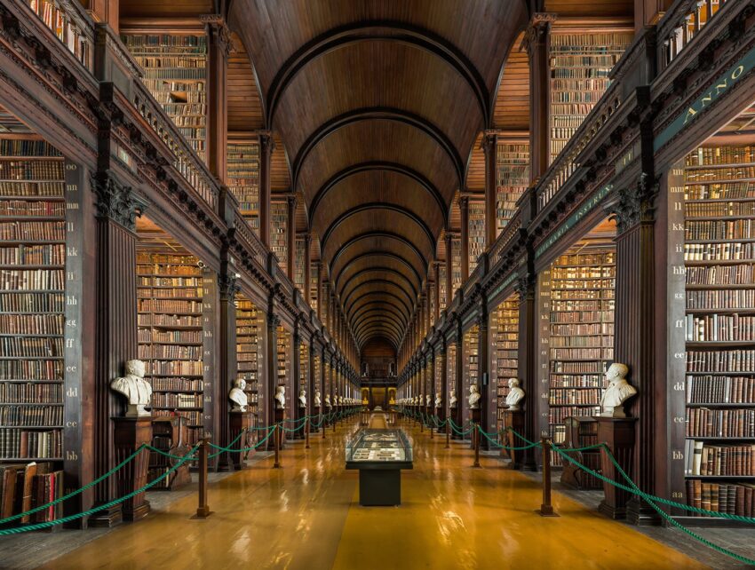 Interior view of a historic library's long room with wooden arches, bookshelves, and statues lining the corridor.