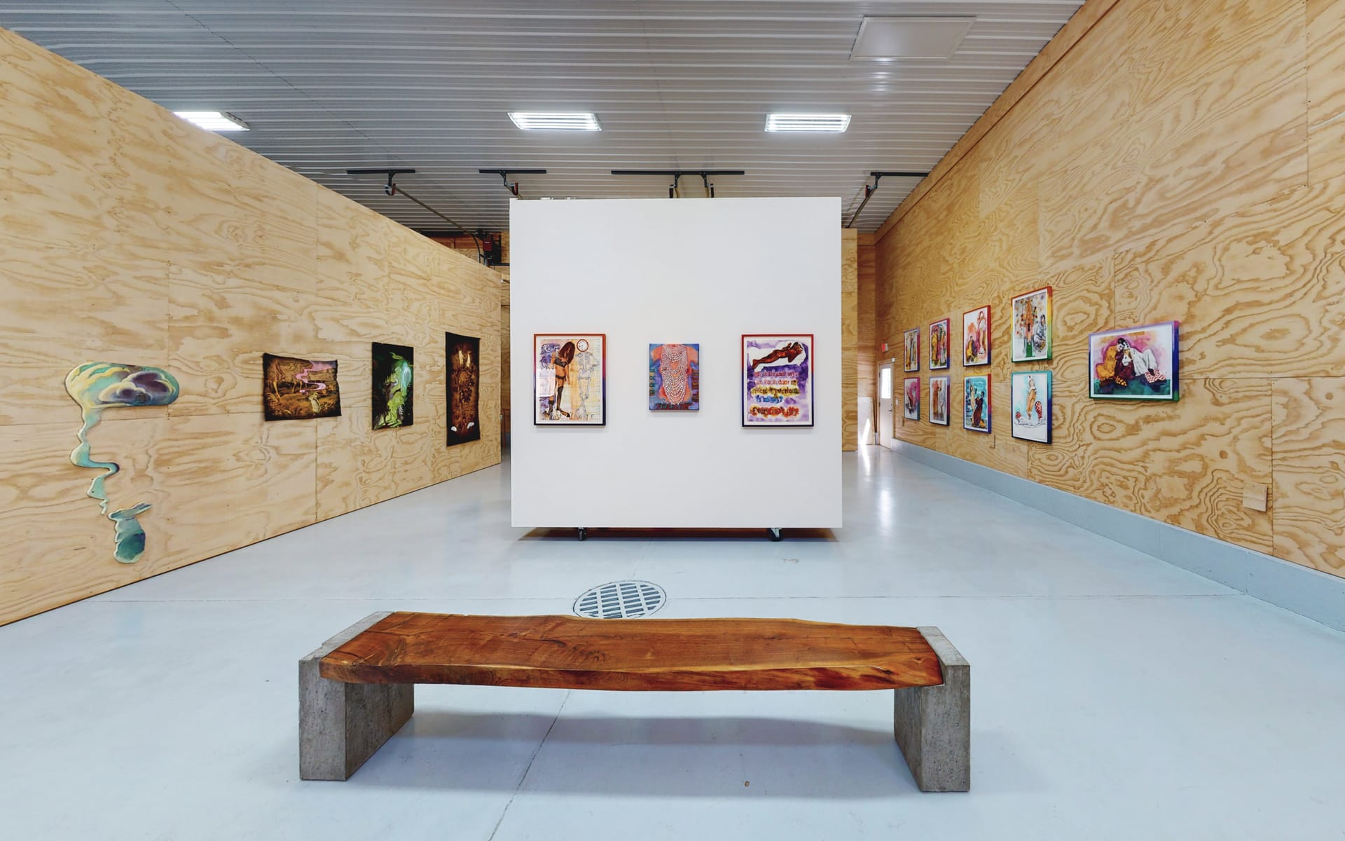 Art gallery interior with wooden walls, a variety of colorful paintings on display, and a wooden bench in the foreground.