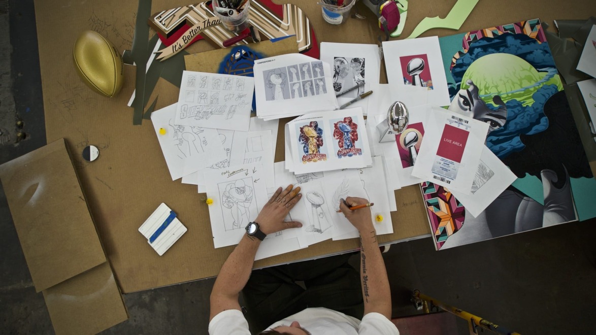 Artist sketching on a cluttered desk filled with drawings, colorful artwork, and art supplies from a bird's-eye view.