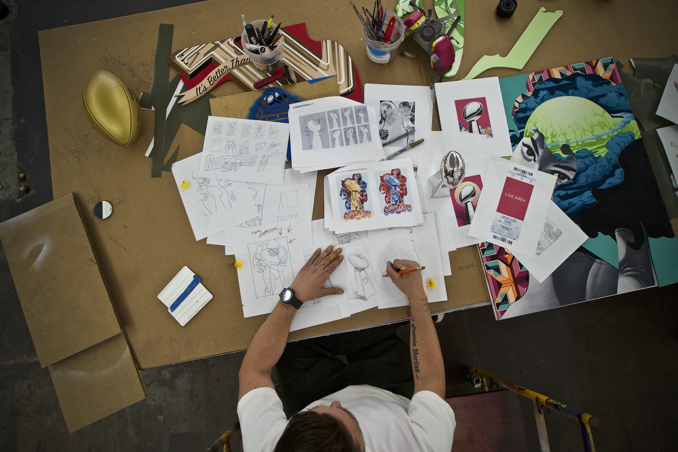 Artist sketching on a cluttered desk filled with drawings, colorful artwork, and art supplies from a bird's-eye view.