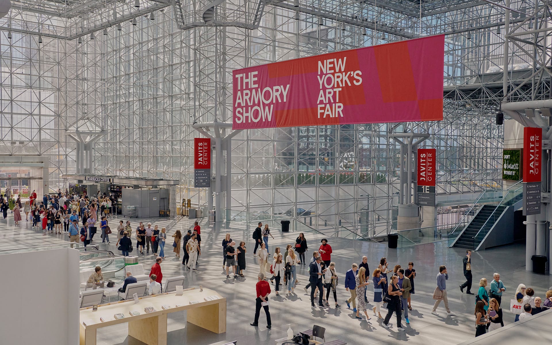 People walking inside a large exhibition hall for New York's Armory Show art fair, with colorful banners overhead.