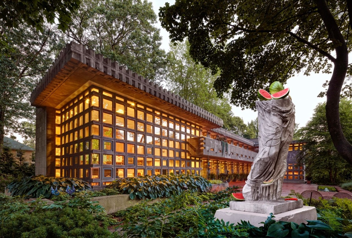Modern building with illuminated windows surrounded by trees and a humorous statue with watermelon slice details in the foreground.
