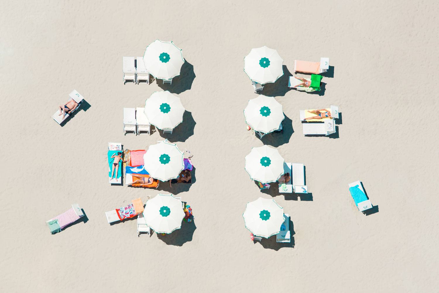Aerial view of a sandy beach with white umbrellas and people sunbathing on lounge chairs.