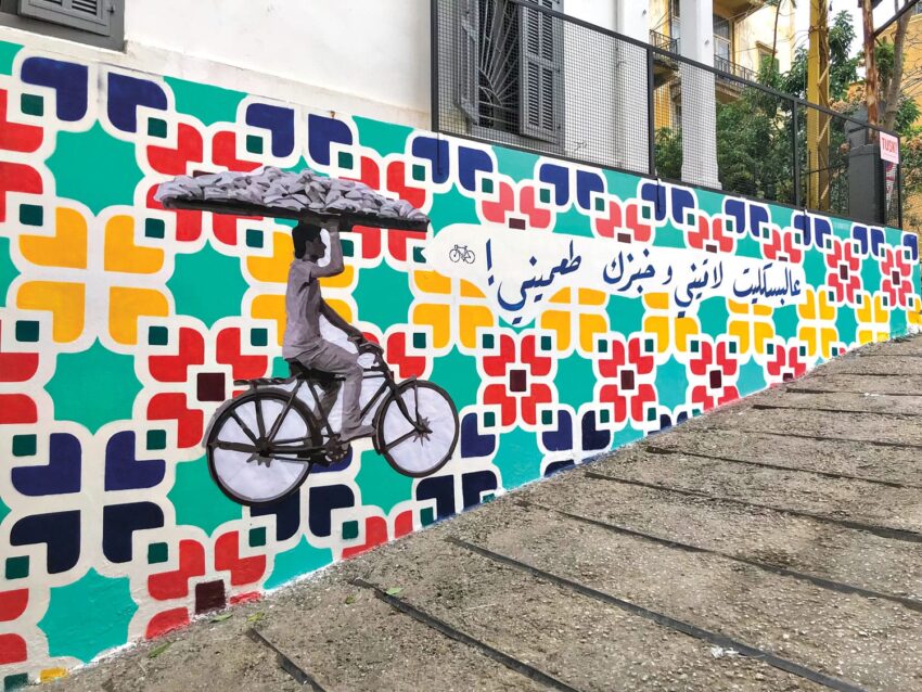 Colorful mural of a cyclist with a tray of bread against a vibrant geometric pattern and Arabic script on a sloped street wall.