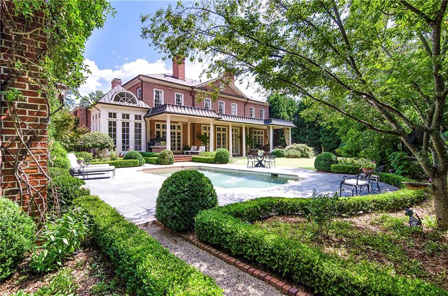 Elegant brick house with large windows, surrounded by lush greenery, featuring a patio area and a serene rectangular pool.