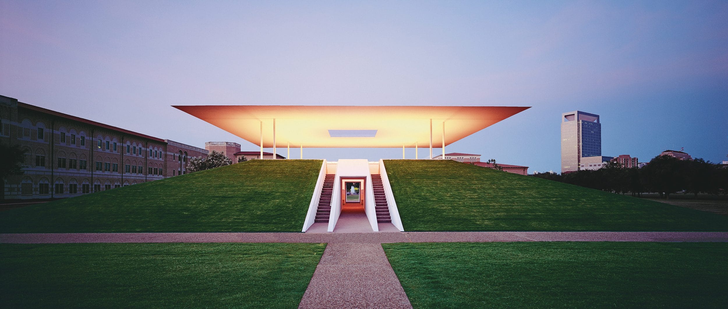 Illuminated modern pavilion atop a grass mound with stairs and a cityscape background at dusk.