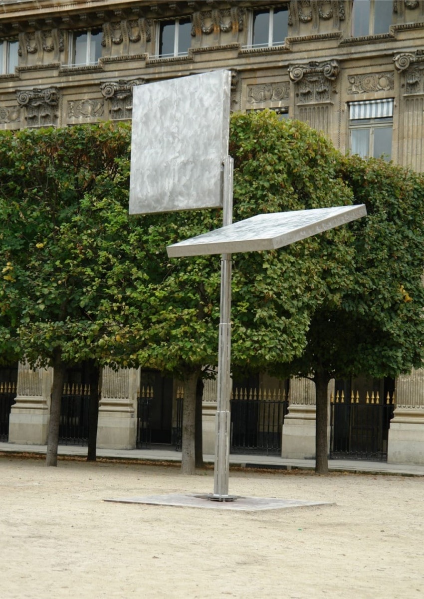 Modern outdoor metal sculpture with geometric shapes set against a backdrop of trees and a historic building.