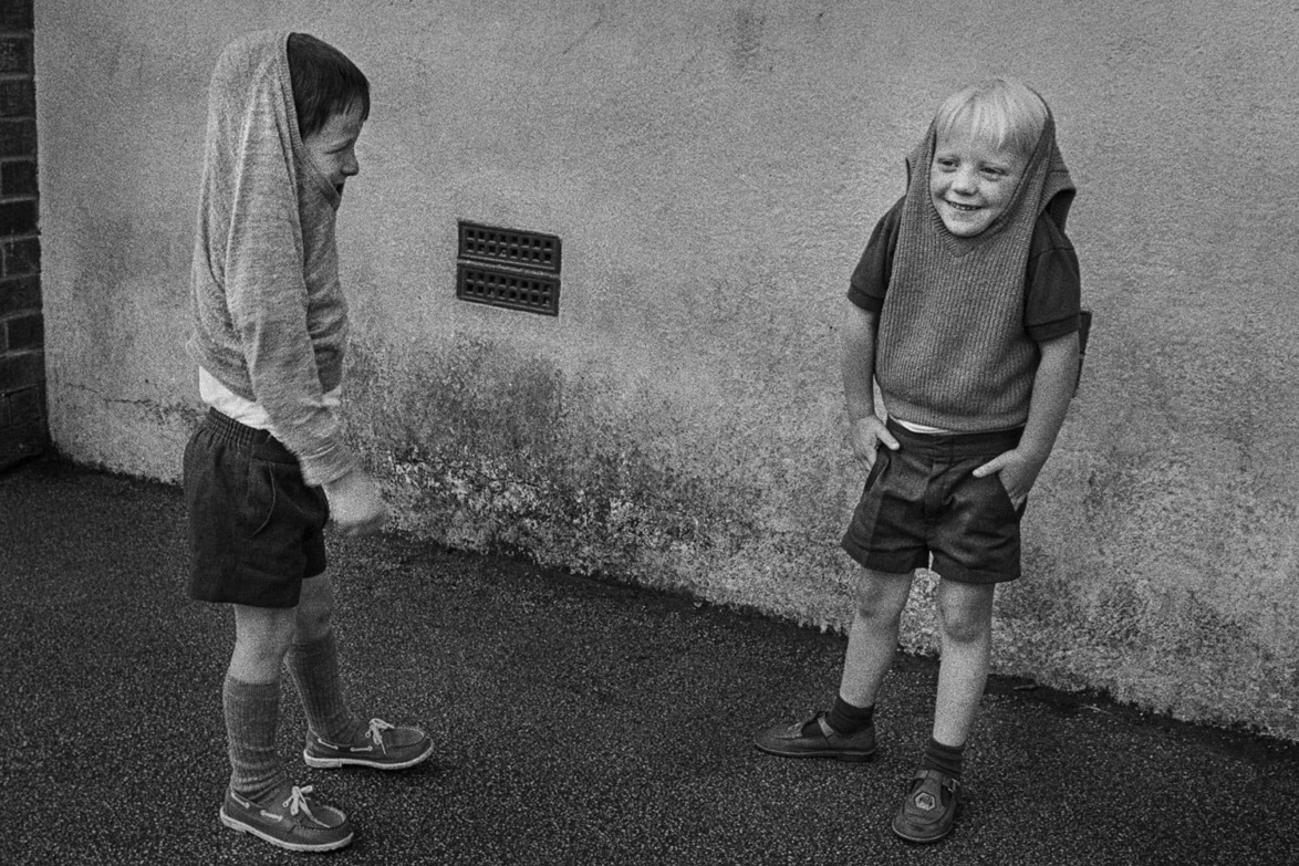 Two children wearing hoodies playfully with hoods up, standing outside on a paved area against a wall.