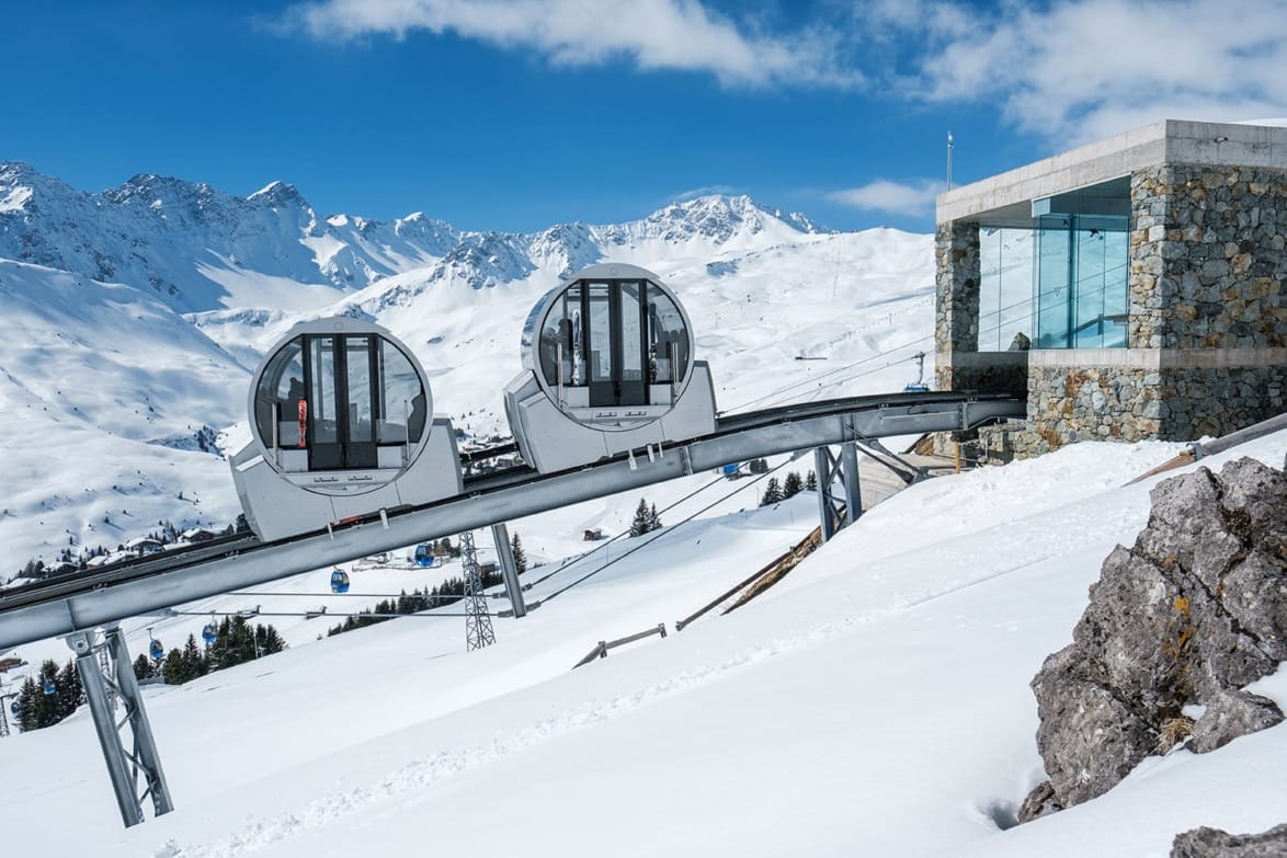 Mountain tram with glass gondolas on tracks, snowy peaks in the background, modern stone and glass station building.