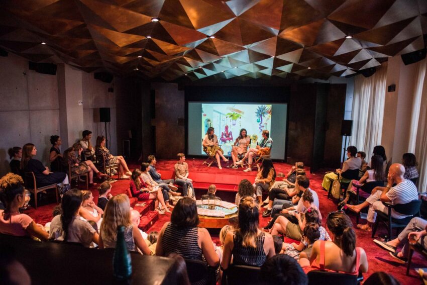 A group of people seated in a room with a panel discussion happening at the front and a large screen displaying visuals.