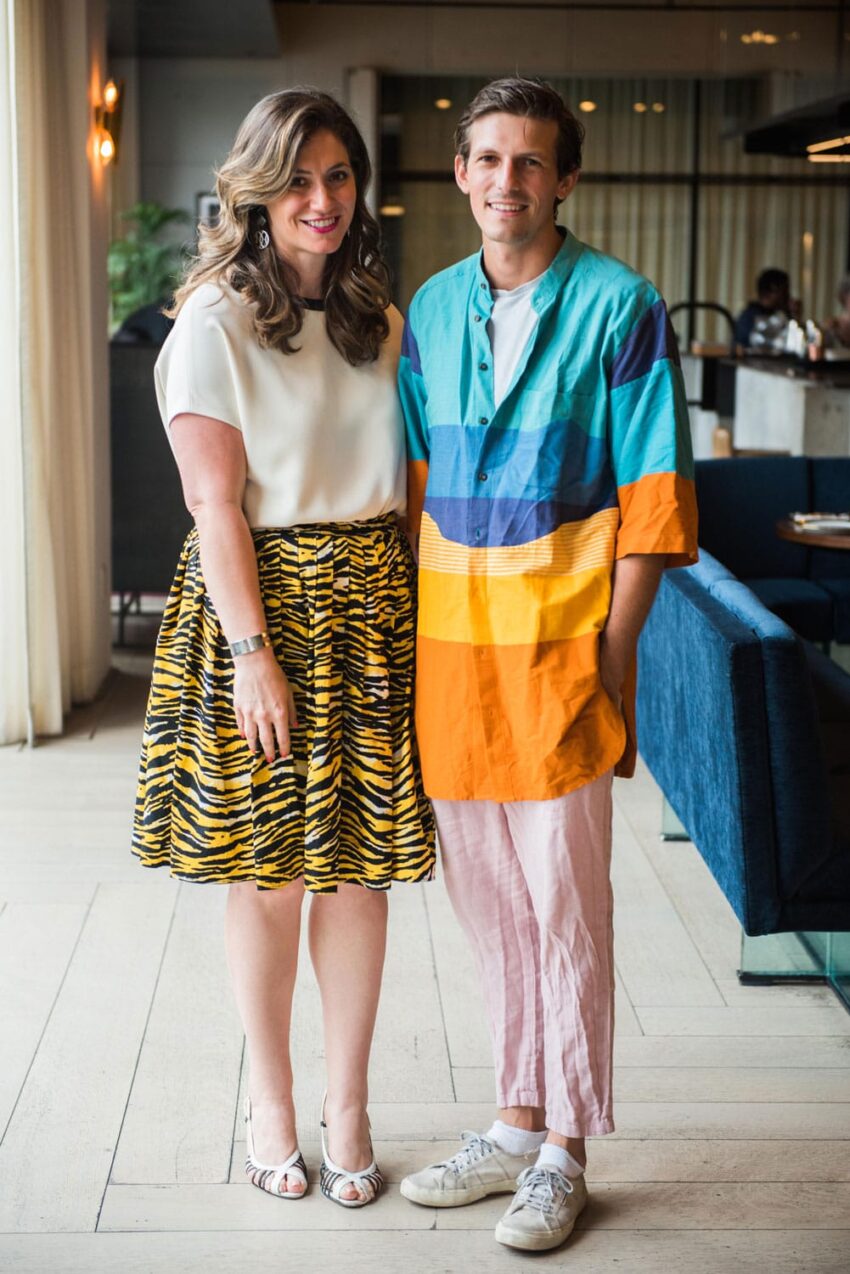 A smiling couple stands in a room with blue chairs, the woman wearing a patterned skirt and the man in a colorful shirt.
