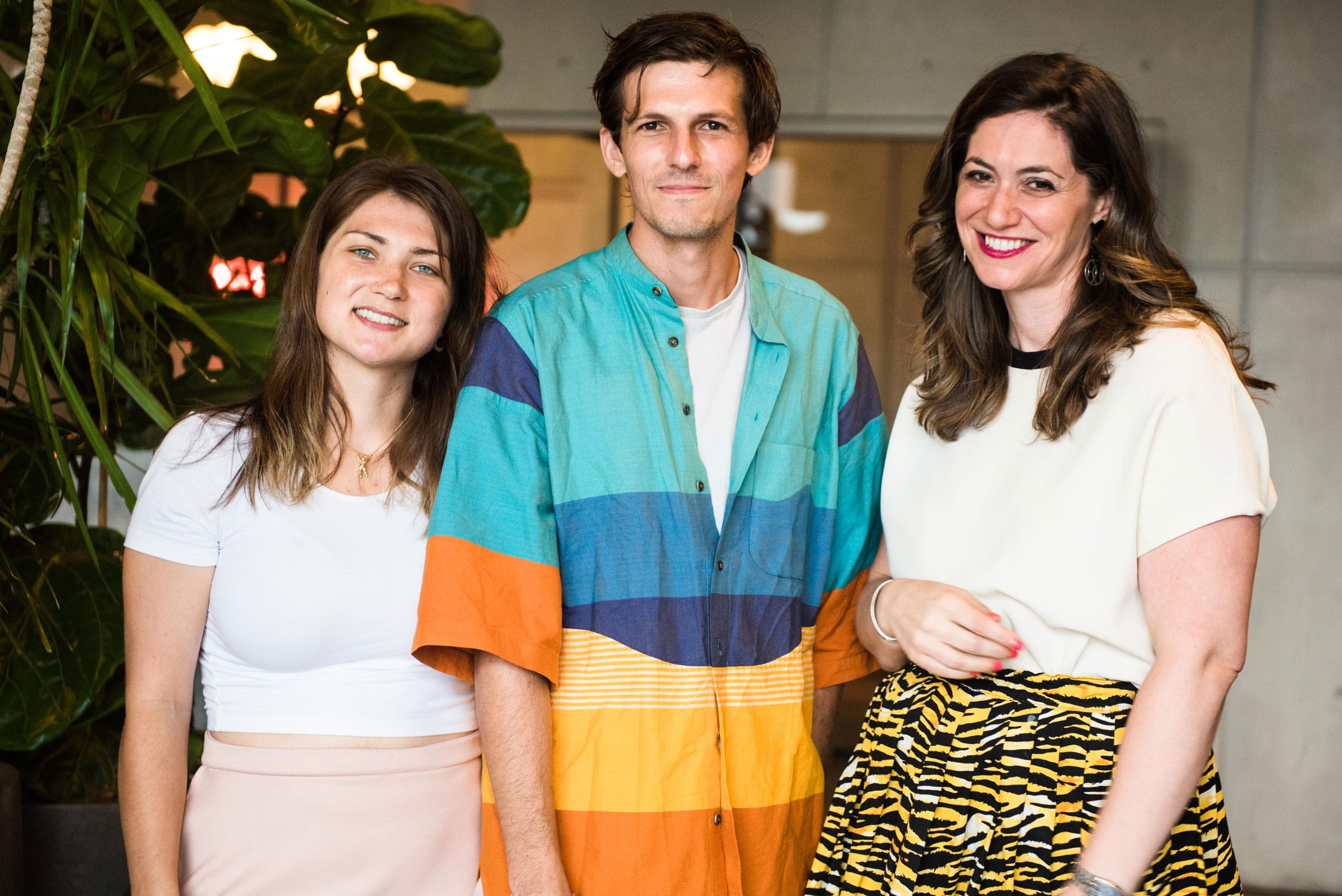 Three people standing together and smiling, with greenery in the background.