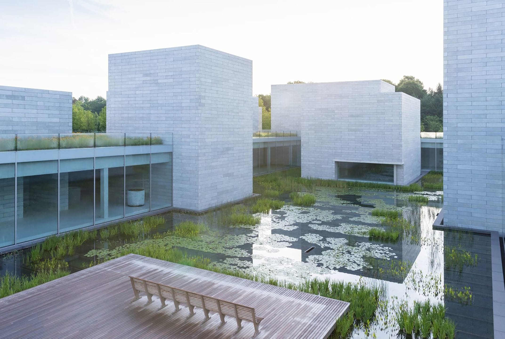 Modern building complex with reflective water features and a wooden deck, surrounded by greenery on a sunny day.