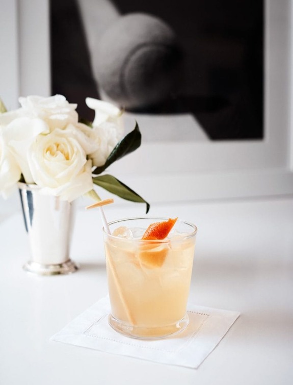 Cocktail with orange garnish on table, next to white roses in vase and framed black and white photo in background.