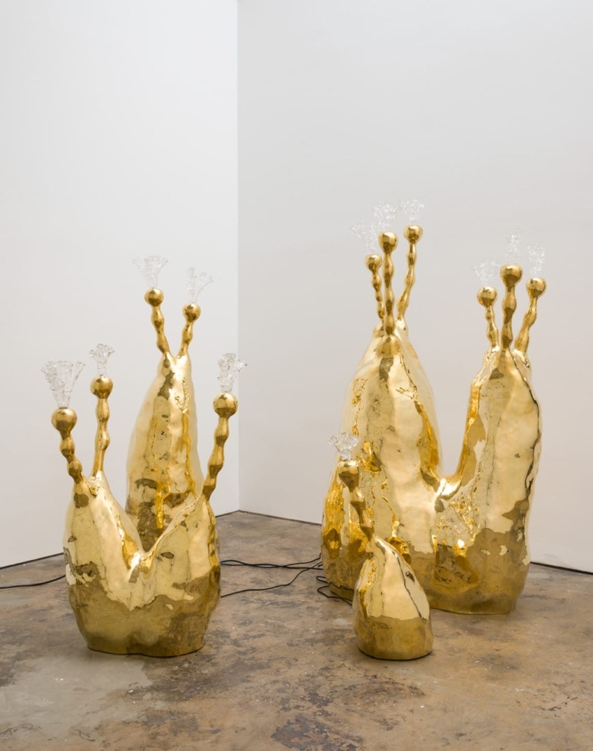 Gold abstract sculptures resembling organic forms with crystal-like accents, displayed in a corner on a concrete floor.