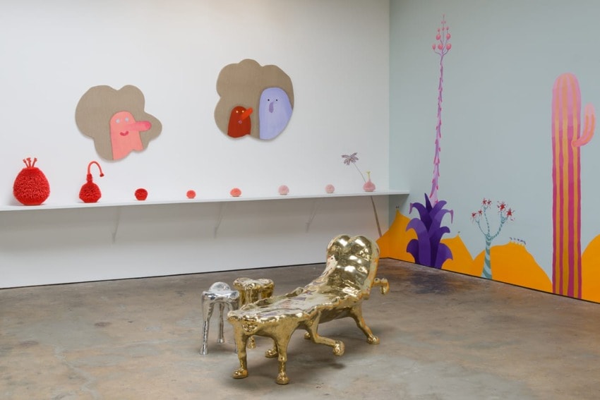 Art gallery interior with quirky gold furniture, colorful wall paintings, and whimsical artwork on display shelves.