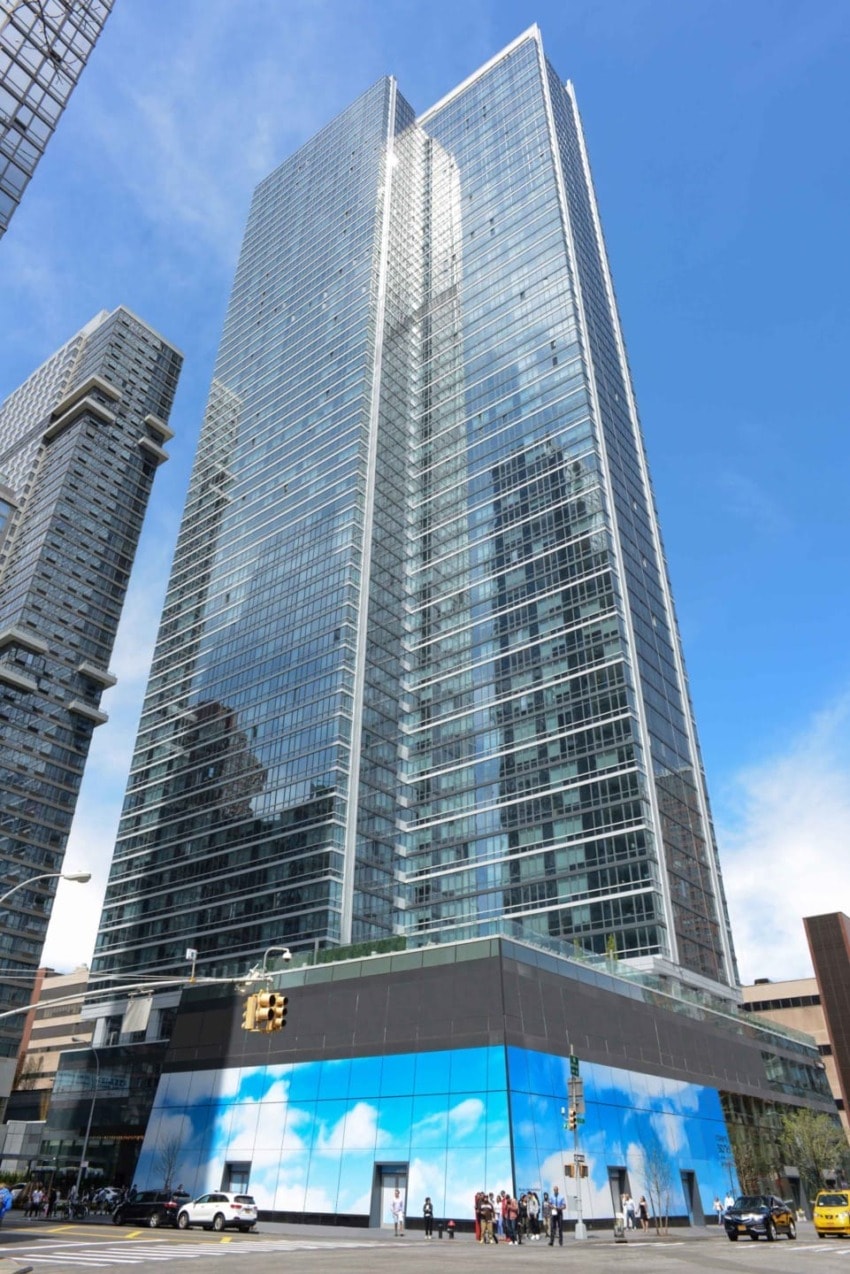 Modern skyscraper with glass facade against a blue sky, street-level view with pedestrians and traffic signal in foreground.