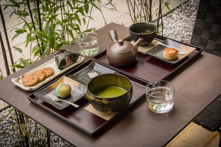 Traditional Japanese tea set with green tea, teapot, sweets, and rice ball on a wooden table outdoors surrounded by plants.