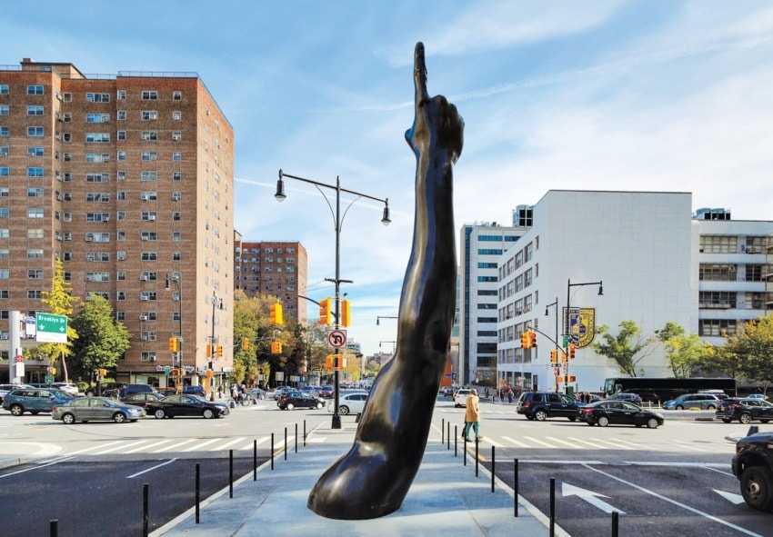 Bronze sculpture of an elongated arm pointing up, set in an urban cityscape with buildings and busy streets in the background.