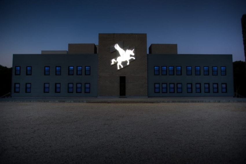 Building facade with illuminated pegasus silhouette at night, surrounded by dark sky and gravel foreground.