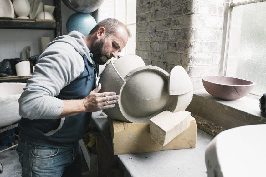Man shaping a large ceramic pot in a workshop, surrounded by pottery tools and materials.