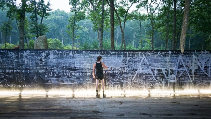 Person writing on a large outdoor chalkboard surrounded by trees and natural scenery.