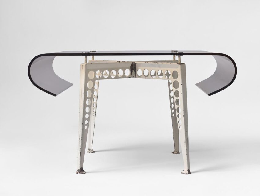 Modern desk with a clear glass top, flared sides, and perforated metal legs, set against a white background.