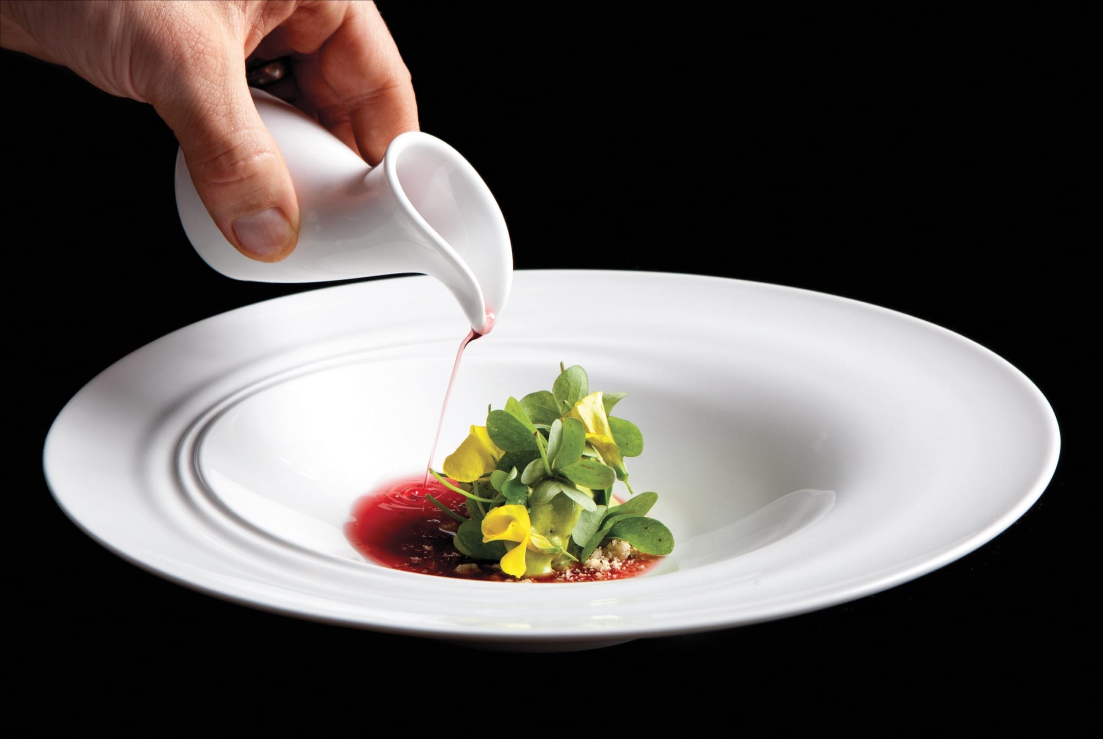 Hand pouring sauce onto a gourmet dish with yellow flowers and greens on a white plate against a black background.