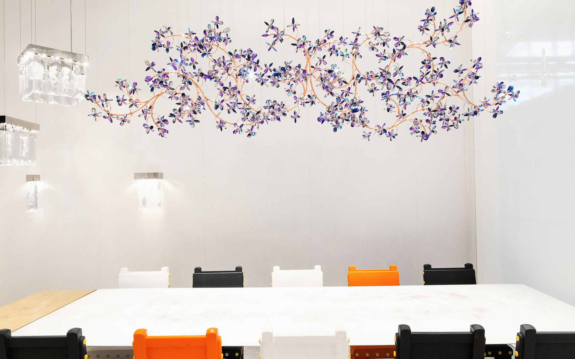 Modern dining room with a white table, colorful chairs, and a floral ceiling light fixture against a white wall.